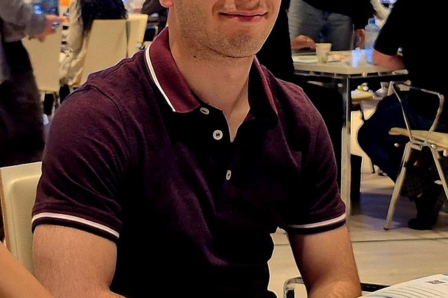 Ein Schüler sitzt an einem Tisch und lächelt in die Kamera. Vor ihm liegt das Businessmaster-Brettspiel mit Spielfiguren, Spielkarten und Spielgeld in verschiedenen Farben. Im Hintergrund sind weitere Teilnehmer*innen an Tischen zu sehen, die ebenfalls am Wettbewerb teilnehmen. Die Atmosphäre wirkt lebendig und konzentriert.